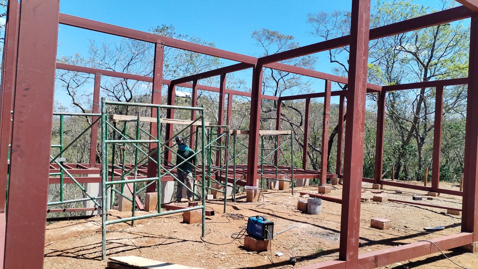 Se observa la estructura metálica de una construcción en proceso con un trabajador y andamios en el sitio.